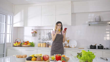 Young asian woman drinking juice near salad table while Young woman watching cellphone in kitchen at home. Health food and technology. Healthy nutrition salad concept for foodie lover.の写真素材