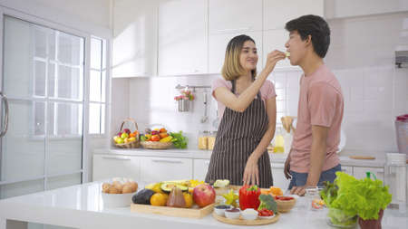 Asian young couple cooking dinner together, cutting fresh vegetables on in kitchen. Woman giving slice of food to her boyfriend to taste. They are enjoy together.Family cooking relationship concept.の写真素材