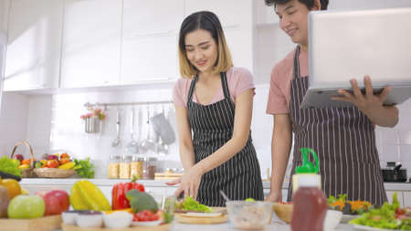 Young asian woman eating sandwich breakfast making roasted bread on electric toaster. Young man watching laptop preparing meal together with affection and love. Health food and technology.の写真素材