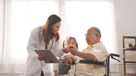 Asian female nurse tells how to take medicine to elderly senior grandfather patient at home medical visit, Elderly old male people healthcare support and encouraging the patient with granddaughter.の写真素材