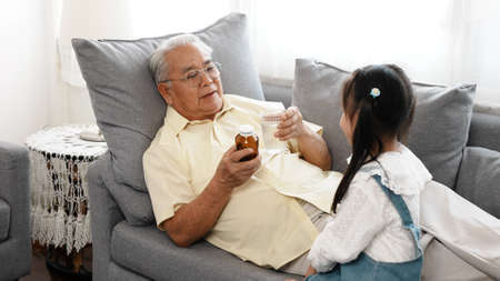 Happy family Little asian daughter carried a drug and glass of water to the sick grandfather on the sofa in the house take care, Elderly old man patient retired enjoy life concept.の写真素材