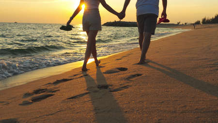 Couple walking together on the beach at sunset, young happy couple holding hands walking along beach, walking barefoot and carrying shoes, outdoor leisure time by the seaside.の写真素材