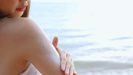 Close up Smiling young asian woman applying a sunscreen lotion on shoulder her skin on a seaside...