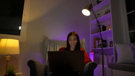 Happy young asian woman using laptop teachnology sitting on armchair relaxing after work in living room at night.の写真素材