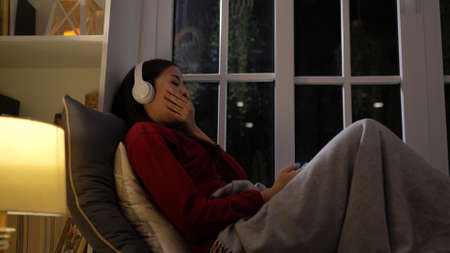 Happy young woman listening music and yawning on sofa after hard working in living room at night, Cozy relaxing at home concept.の写真素材