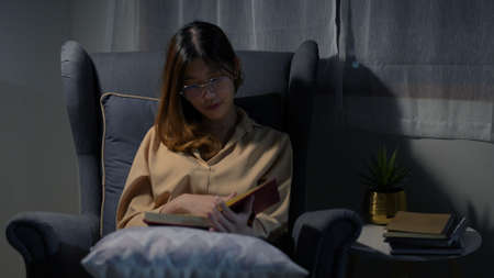 Happy young asian woman reading book sitting on sofa relaxing after work in living room at night.の写真素材