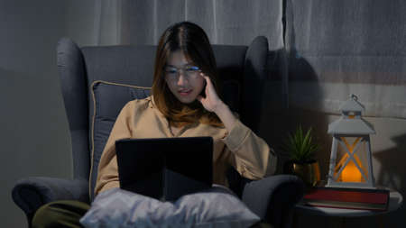 Happy young asian woman reading book sitting on armchair relaxing after work in living room at night.の写真素材