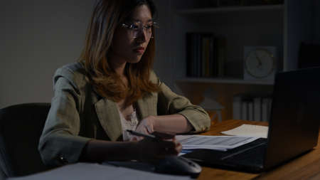 Asian businesswoman or freelancer working hard at the night looking the laptop digital tablet and document in workplace at late overtime deadline, Stressed busy and exhausted of work at home.の写真素材