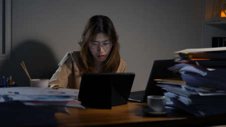 Asian businesswoman or freelancer working hard at the night looking the laptop digital tablet and document in workplace at late overtime deadline, Stressed busy and exhausted of work at home.の写真素材