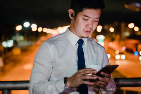 Asian businessman use mobile phone, smart phone after work walking through city street while waiting car in the city at nightの写真素材