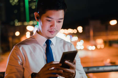 Asian businessman use mobile phone, smart phone after work walking through city street while waiting car in the city at nightの写真素材
