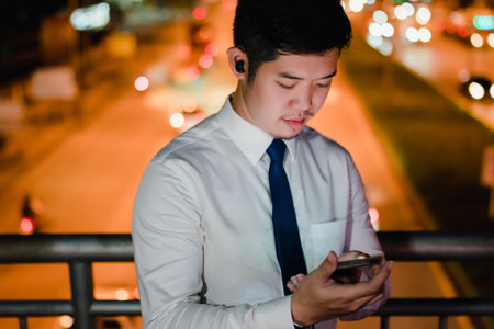 Asian businessman use mobile phone, smart phone after work walking through city street while waiting car in the city at nightの写真素材