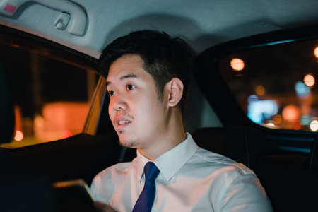 Asian businessman use mobile phone, smart phone after work walking through city street while waiting car in the city at nightの写真素材