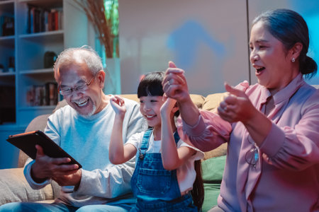 Asian happy family stay home in living room at night. Little girl with grandparents dancing having fun together on sofa. Party laughing, enjoy parenting activity relationship in house.の写真素材