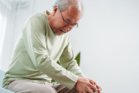 Asian Senior man feel knee pain ache. Older mature male hurt at knee while sitting on sofa in living room at home. Bone problam health care and medical insurance concept.の写真素材
