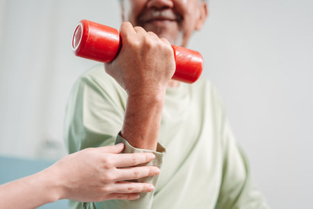 Asian Senior old man doing physiotherapist with support from caregiver. Mature male sitting on sofa in living room using dumbbell workout exercise. Healthcare medical concept.の写真素材