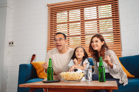 Asian Family with daughter watching sport football cheering excited eating popcorn on sofa in living room at home. enjoy relaxing happiness.の写真素材