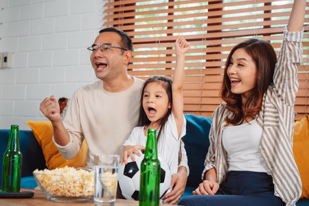 Asian Family with daughter watching sport football cheering excited eating popcorn on sofa in living room at home. enjoy relaxing happiness.の写真素材