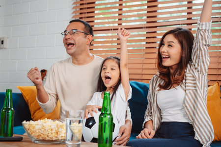 Asian Family with daughter watching sport football cheering excited eating popcorn on sofa in living room at home. enjoy relaxing happiness.の写真素材