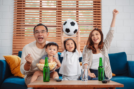 Asian Family with daughter watching sport football cheering excited eating popcorn on sofa in living room at home. enjoy relaxing happiness.の写真素材