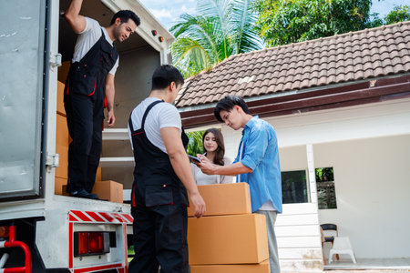 Asian Couple check while unloading boxes and furniture from a pickup truck to a new house with service cargo two men movers worker in uniform lifting boxes. concept of Home moving and delivery.の写真素材