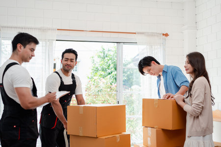 Asian Couple check while unloading boxes and furniture from a pickup truck to a new house with service cargo two men movers worker in uniform lifting boxes. concept of Home moving and delivery.の写真素材