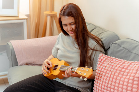 Woman Asian Playing Music Ukulele Lifestyle Relax Lesson Fun Relaxation, Cheerful Moment of Home Ukulele Practice, Sharing Joy and Music in a Cozy Living Room with Acousticの写真素材