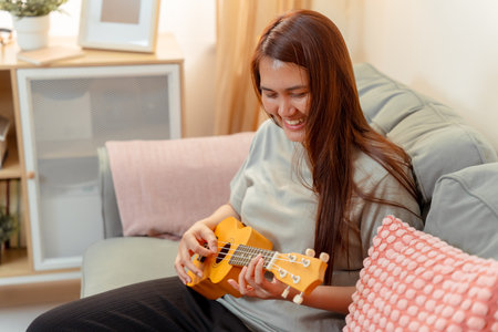 Woman Asian Playing Music Ukulele Lifestyle Relax Lesson Fun Relaxation, Cheerful Moment of Home Ukulele Practice, Sharing Joy and Music in a Cozy Living Room with Acousticの写真素材