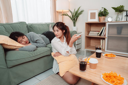 Asian Women Snack Food Eating Lifestyle Indoor Living Room Enjoyment Movie, Two Young Adults Sharing Laughter and Snacks While Watching TV, Embracing Friendship and Relaxation at Home Togetherの写真素材