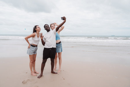Diverse group of young friends taking selfie and enjoying time together on the beach. Fun summer vacation moment symbolizing friendship, freedom, and multicultural connection.の写真素材