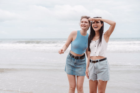 Two young women friends smiling and looking out at the ocean while standing on the beach. Perfect summer vacation vibe showing friendship, fun, freedom, and joyful outdoor lifestyle.の写真素材