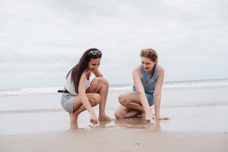 Two young women sitting on the beach drawing on sand, enjoying fun and creative moments during...