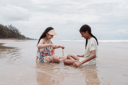 Multicultural and Asian daughters sitting together on the beach, smiling and enjoying a peaceful moment of friendship, childhood joy, diversity, and seaside happiness.の写真素材