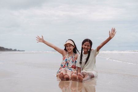 Asian and multicultural daughters sitting with smiles and raised hands on the beach, childhood joy, friendship, diversity, freedom, and summer vacation happiness.の写真素材