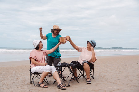 Asian senior females enjoying drinks and laughter together on the beach, retirement togetherness,...