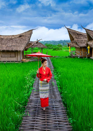 Young Thai woman wearing traditional Lanna clothing walks on a bamboo bridge in the middle of a rice field with a backdrop of a wooden resort in Nan Province, Thailandの写真素材