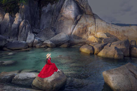 Young woman tourist in red dress sit on Hin Ta Hin Yai with blue sea and sky during summer travel at Koh Samui island, Surat Thani, Thailandの写真素材