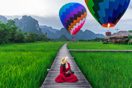 Asian woman holding a camera sits on a wooden walkway and looks at balloons with green rice fields in Vang Vieng, Laos.の写真素材