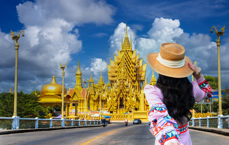A young Asian tourist stands looking at a striking golden temple in Phitsanulok province, Thailandの写真素材