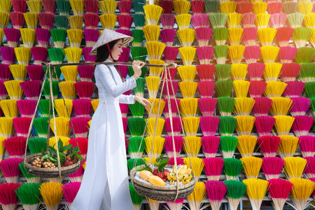 A Vietnamese girl in an ao dai dress carries a fruit basket walking past an incense shopの写真素材