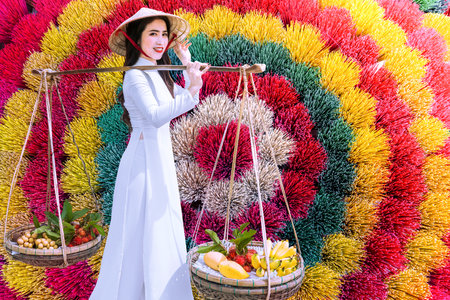 A Vietnamese girl in an ao dai dress carries a fruit basket walking past an incense shopの写真素材
