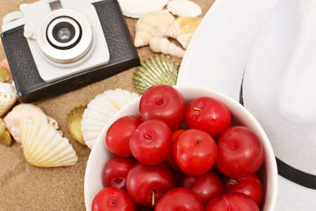 Red plums, old camera, shells and white hat on sandの写真素材