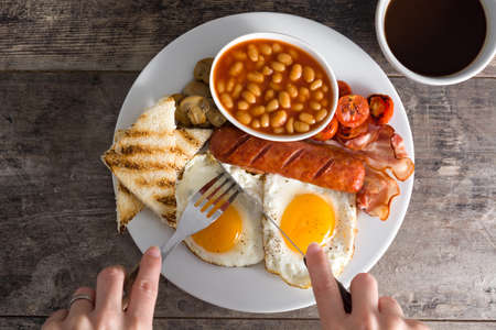 Woman eating a traditional english breakfast with fried eggs, sausages, beans, mushrooms, grilled tomatoes, bacon, coffee and orange juice, on wooden tableの写真素材