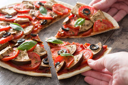 Hands taking vegetarian pizza slice With eggplant, tomato, black olives, oregano and basilの写真素材