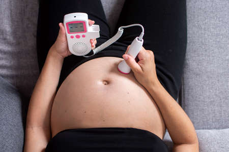 Pregnant woman with a fetal doppler listening to the baby's heart sitting on the sofa at homeの写真素材