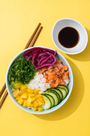 Poke bowl with rice, salmon, cucumber, mango, onion, wakame salad, poppy seeds and sunflower seeds on yellow backgroundの写真素材