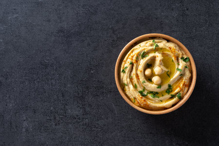 Chickpea hummus in a wooden bowl garnished with parsley, paprika and olive oil on black slate backgroundの写真素材