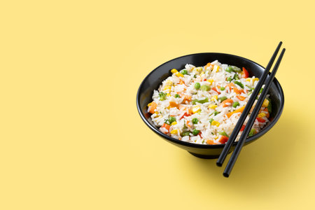 White rice with vegetables in a black bowl on yellow backgroundの写真素材