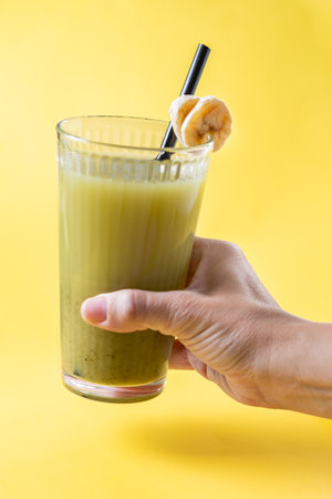 Close-up of a hand holding a tall glass of green smoothie with a black straw and banana slice garnish. The vibrant yellow background gives a fresh and summery feel. A concept of healthy eating, detox drinks, and plant-based nutrition.の写真素材