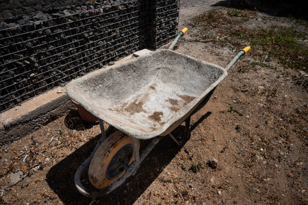 An old, weathered wheelbarrow with a single front wheel stands on dry, dusty soil beside a wire and...